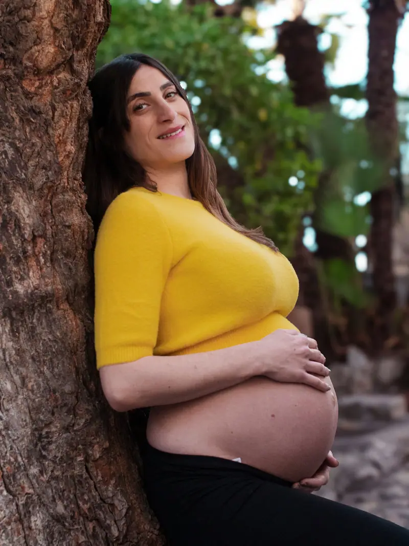 Future maman en pull jaune adossée à un arbre, ventre rond