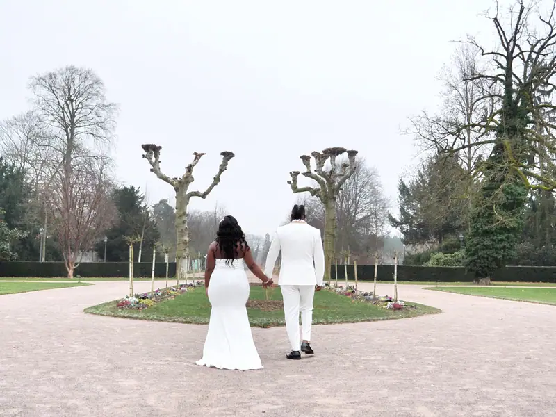 Mariés de dos se tenant la main dans l'allée d'un parc aux arbres taillés