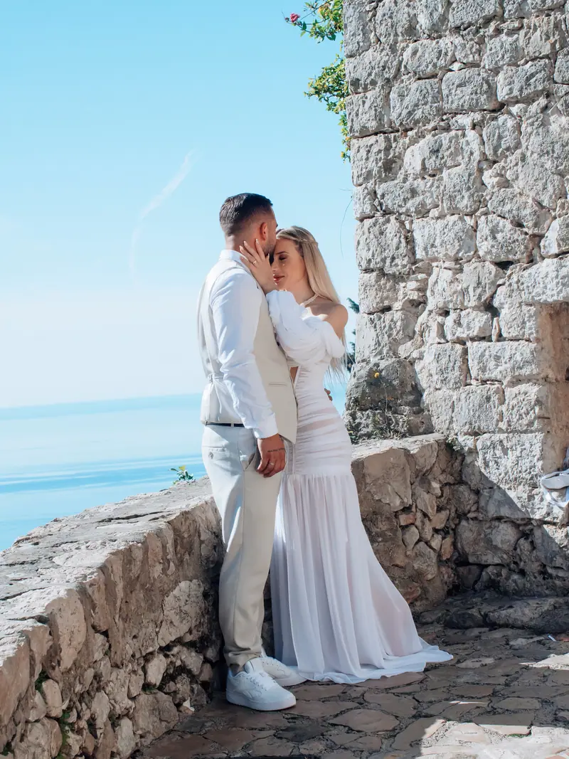 Marié embrassant la mariée sur le front contre un muret en pierre face à la mer