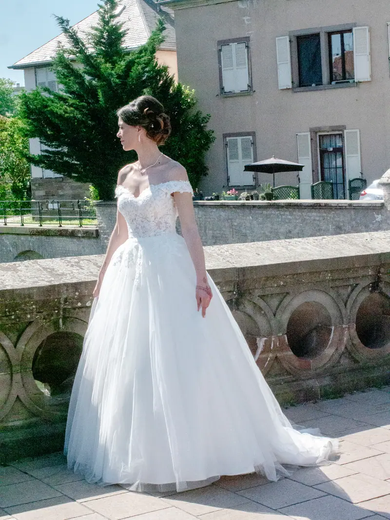 Mariée en robe à traîne posant devant une haie, bâtiments en arrière-plan